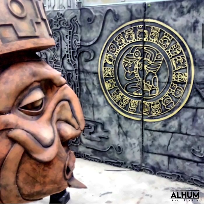 Cabeza monumental tallada frente a un muro con calendario maya en relieve dorado